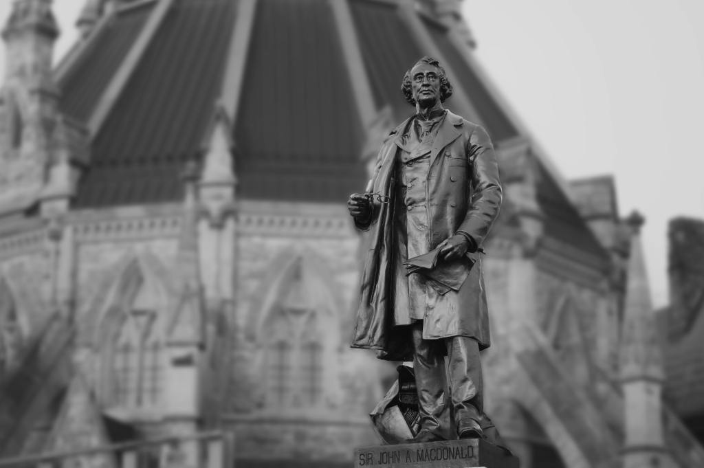 A black and white photo of a statue of Sir John A. MacDonald in the foreground. The Canadian Parliament building looms behind him.