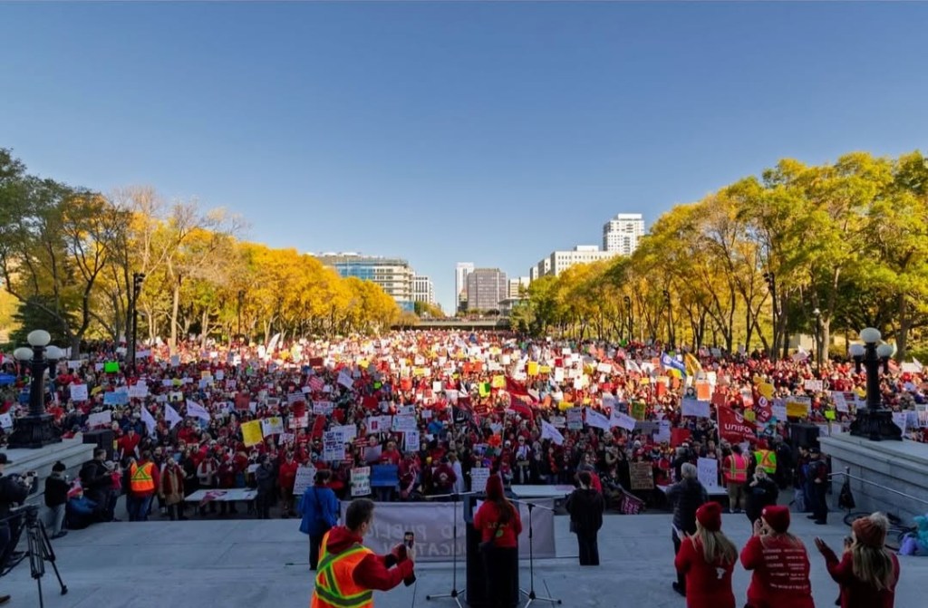 A large crowd of people gathered at an outdoor rally, holding signs and banners, with many participants wearing red attire. Trees with autumn leaves are visible in the background, along with buildings in the distance under a clear blue sky.